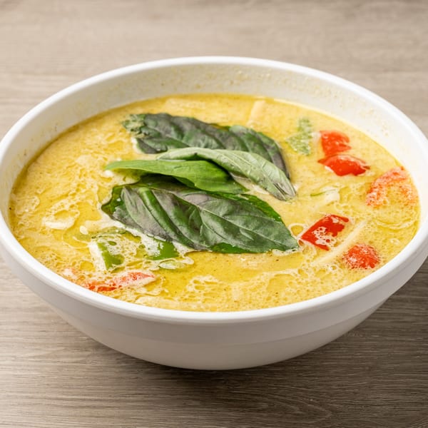 A white bowl filled with vibrant yellow Thai green curry, garnished with fresh basil leaves and red peppers.
