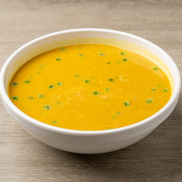 A white bowl filled with vibrant yellow Thai curry, garnished with green peas, on a wooden table.