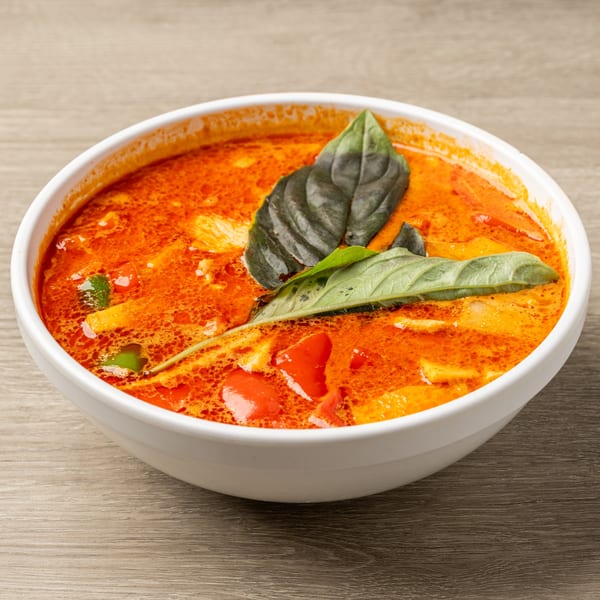 A white bowl of vibrant red Thai curry with vegetables and basil leaves on a wooden table.